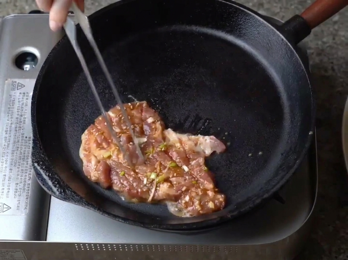 A single piece of seasoned, marinated pork being placed into a preheated black cast iron skillet.
