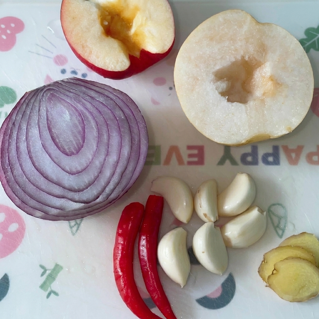 Halved apple, half a red onion, half a pear, garlic cloves, red chilies, and ginger slices arranged on a cutting board.