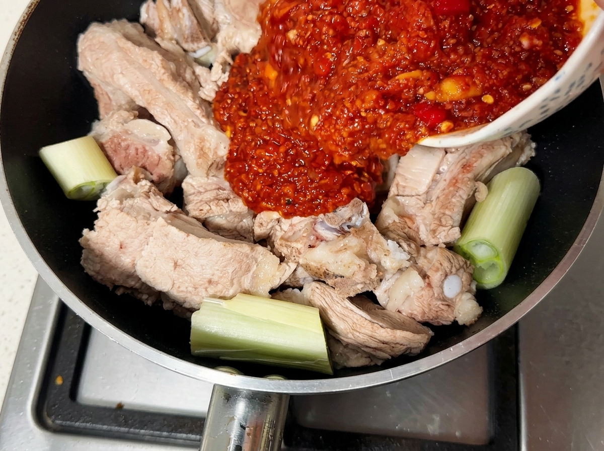 Thick red chili seasoning paste being poured from a bowl over blanched pork ribs in a pan.