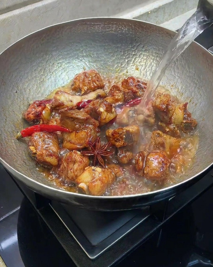 Hot water being poured into a wok containing seared pork ribs and spices.