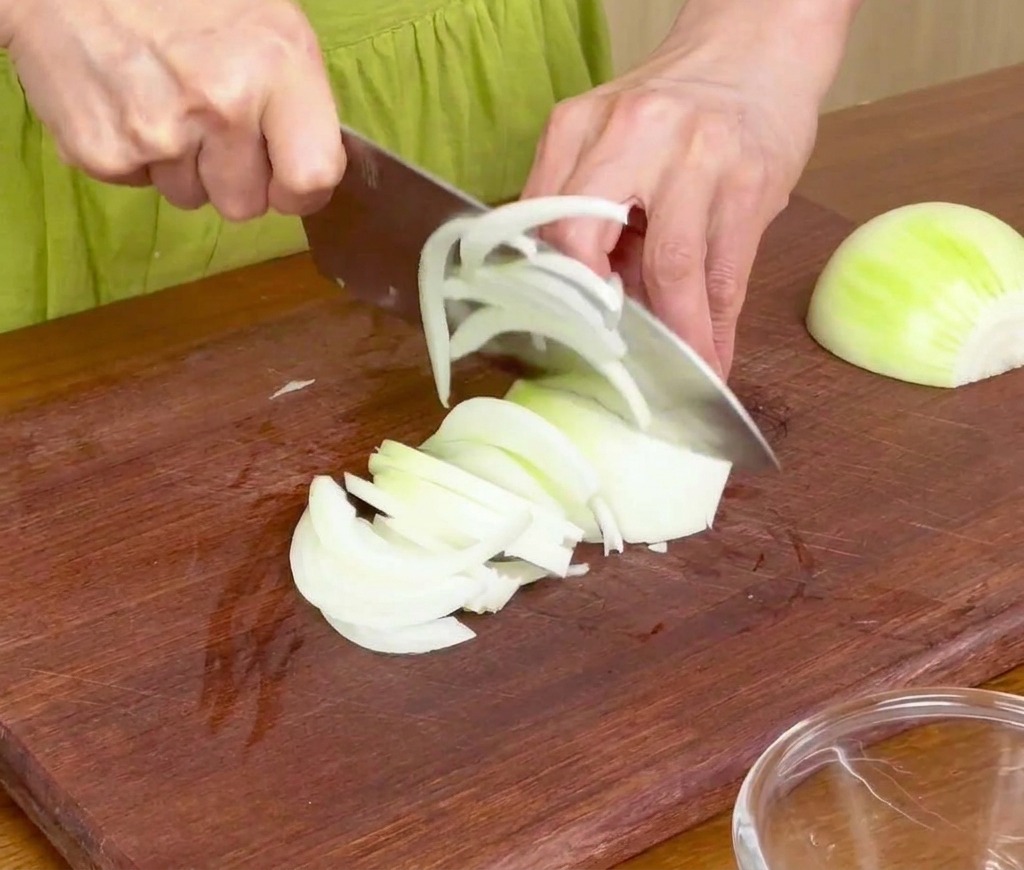 Slicing white onions into thin strips on a wooden cutting board with a chefs knife.