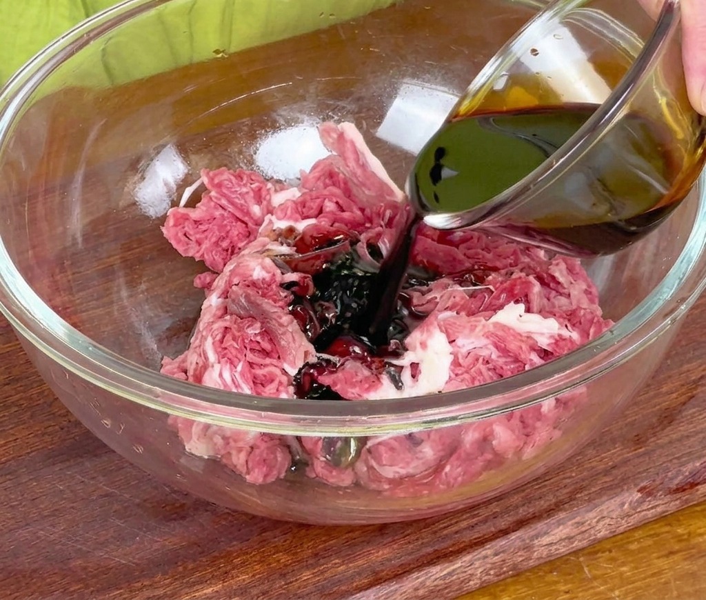 Pouring dark soy sauce from a small glass bowl onto raw sliced beef sitting in a large glass mixing bowl.