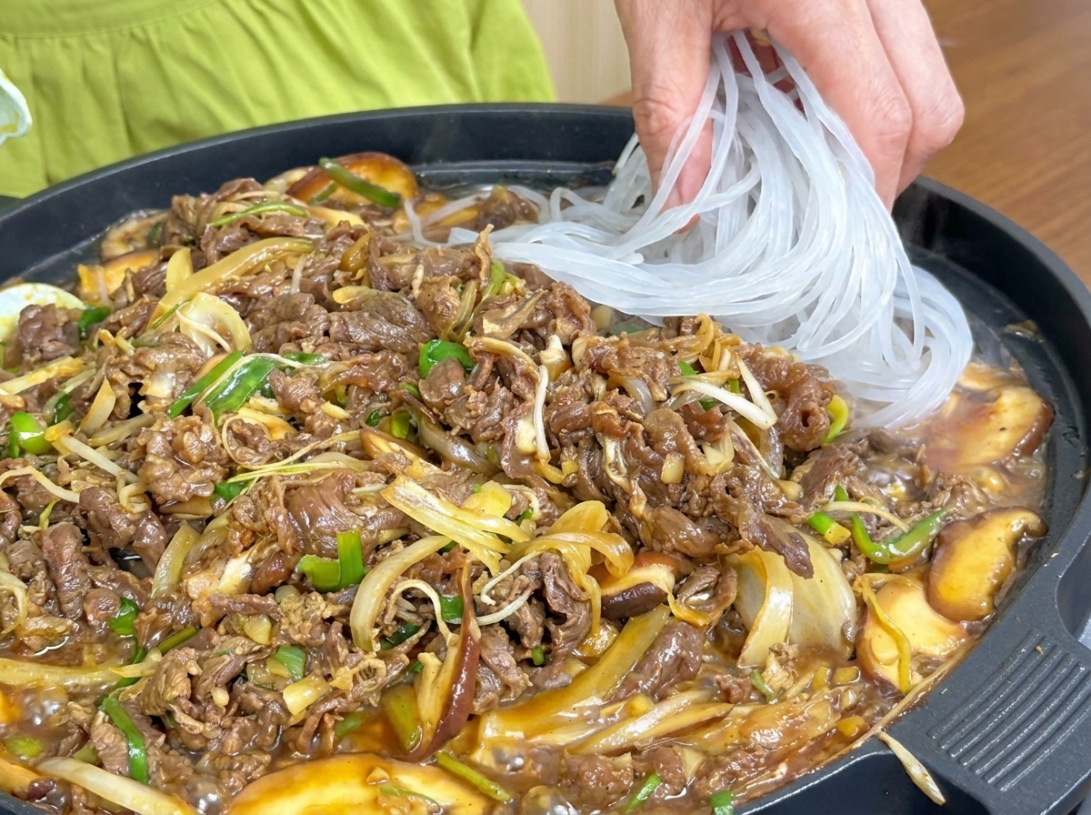 Translucent glass noodles being added to the pan of cooking beef and vegetables.