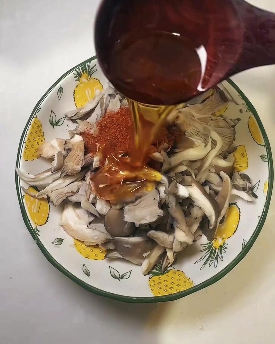 Cooking oil being poured from a wooden ladle over the red chili powder and oyster mushrooms.