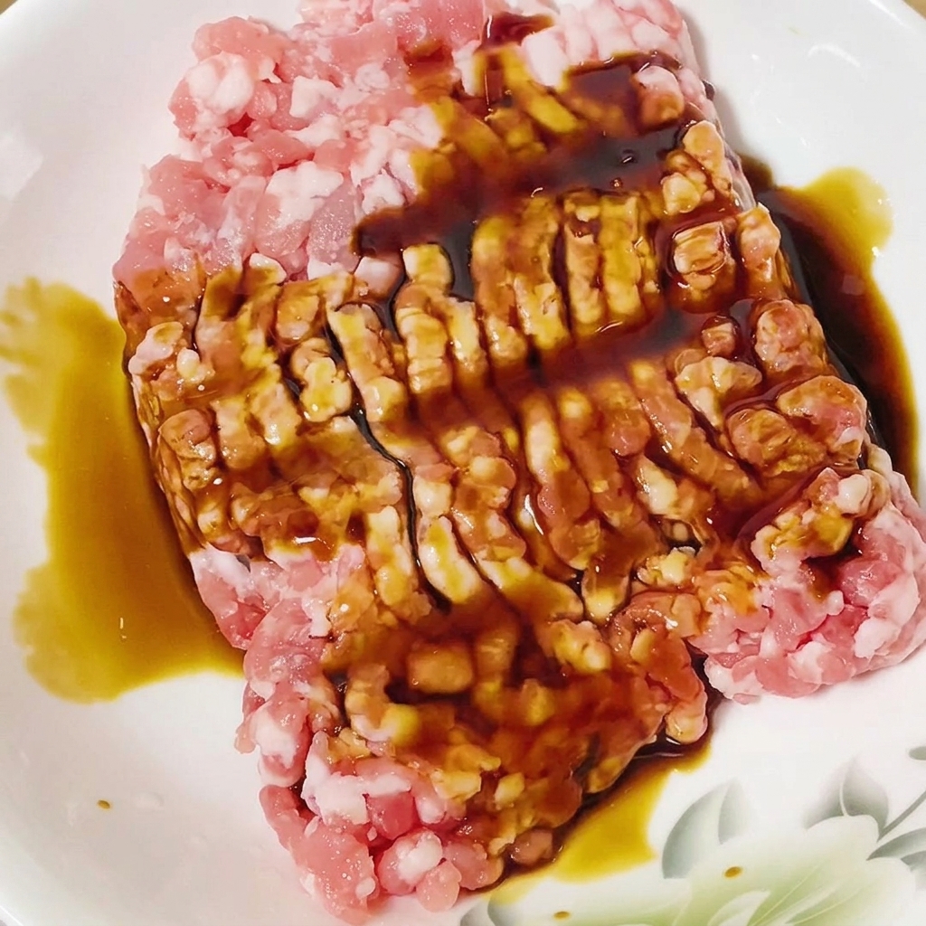 Soy sauce being poured onto raw minced pork in a white bowl.
