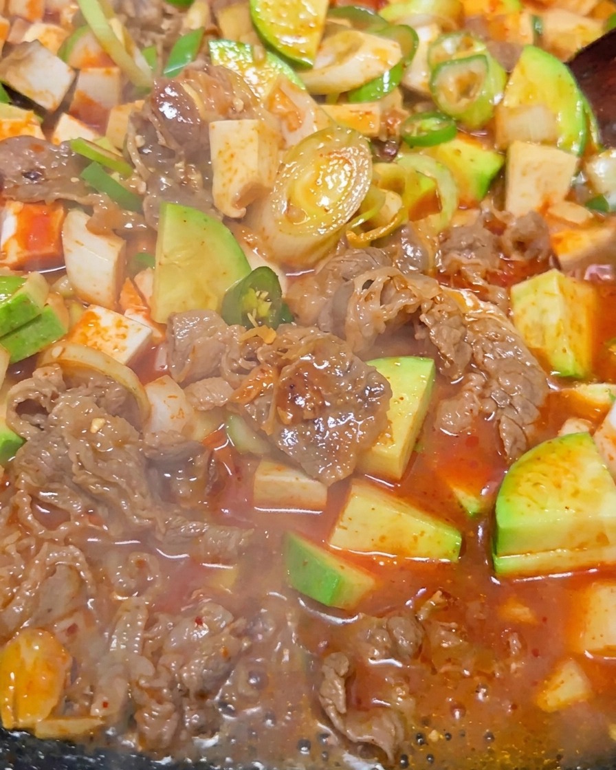 A close-up of the Korean soybean paste stew bubbling intensely, showing chunks of tofu, beef, and green zucchini in a reddish-brown broth.