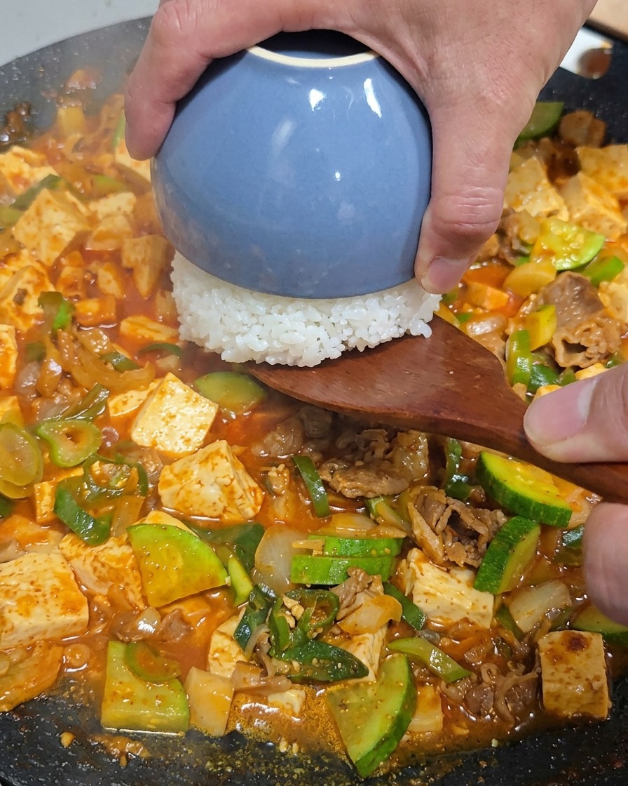 A hand using a wooden spatula to place a small blue bowl of white rice upside down into the center of the pan filled with beef and vegetable stew.