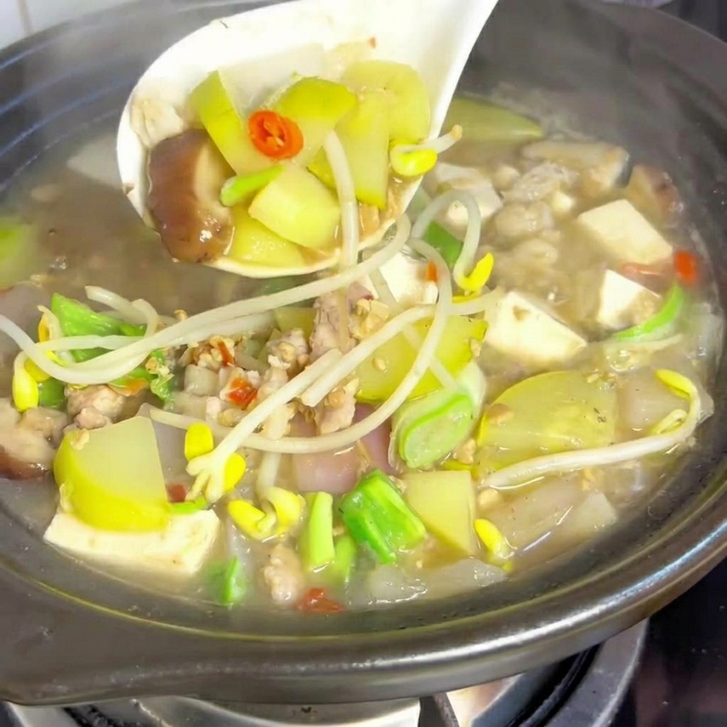 A close-up of a clay pot filled with soybean paste stew, garnished with a handful of fresh yellow soybean sprouts, sliced green onions, and red chili slices.
