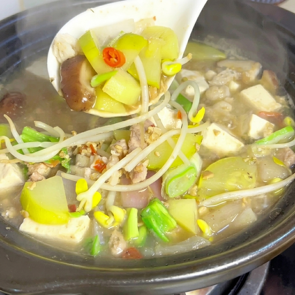 A white ladle gently lifting mixed vegetables, tofu, and meat from a steaming, bubbling clay pot of soybean paste stew.