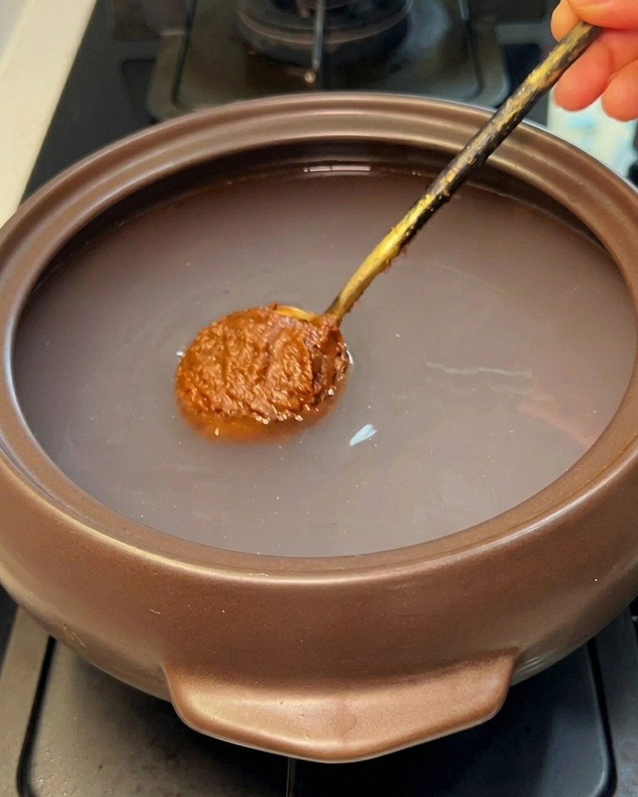 A spoonful of brown Korean soybean paste being submerged into the clear water of a stone pot.