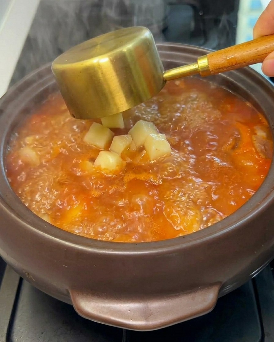 Diced raw potatoes being dropped into the boiling stew from a ladle.