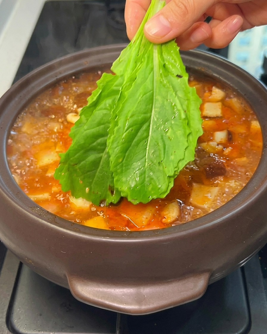 A hand lowering fresh green leafy vegetables into a boiling Korean stew.