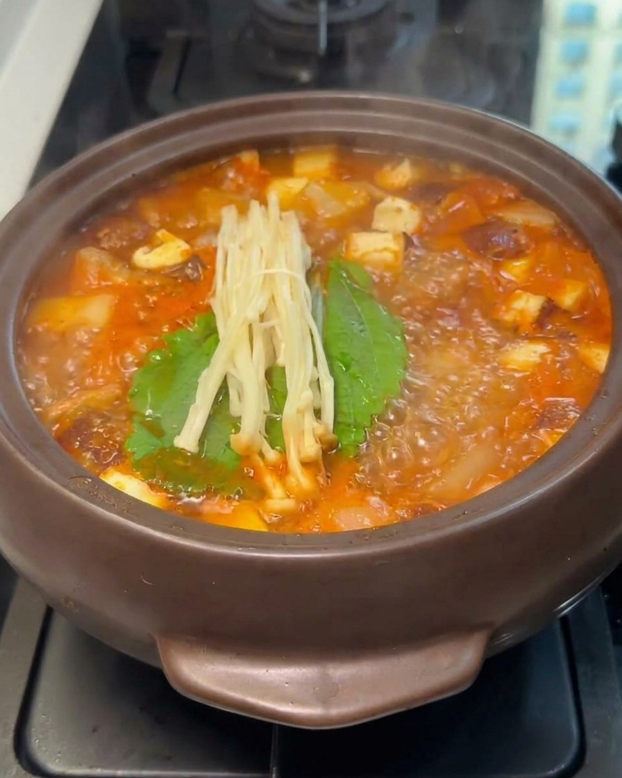 A bundle of fresh enoki mushrooms resting on top of green leaves inside a boiling pot of red stew.