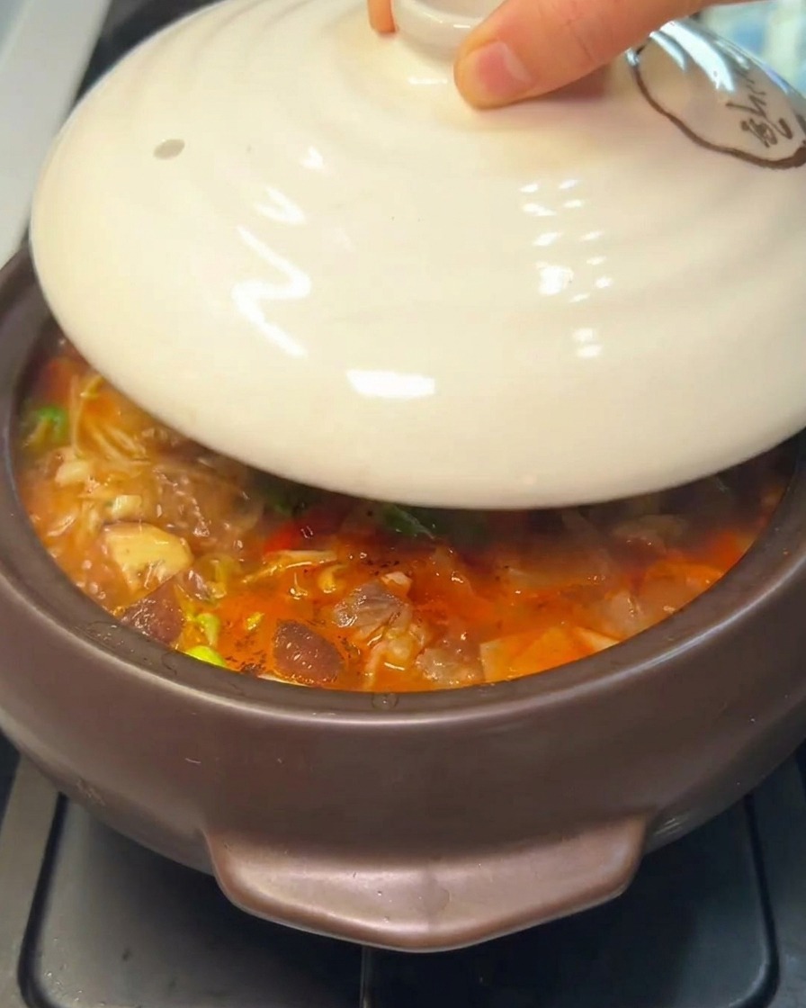 A hand placing a white ceramic lid onto a steaming stone pot of Korean stew.
