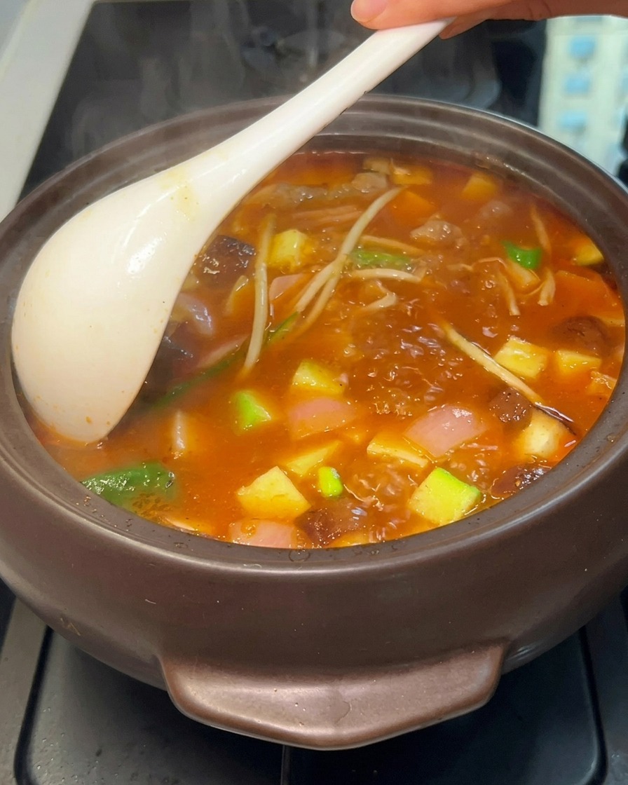 A close-up of a vigorously boiling Korean stew in a dark pot, being stirred with a white ladle to reveal tofu, zucchini, mushrooms, and meat in an opaque, red-orange broth.