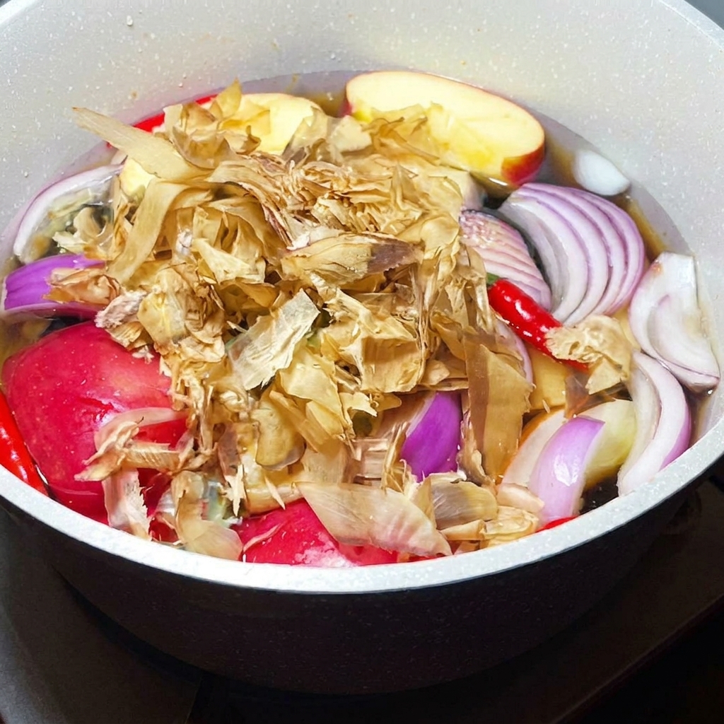Dried bonito flakes being added to a simmering pot filled with aromatic vegetables and fruit.