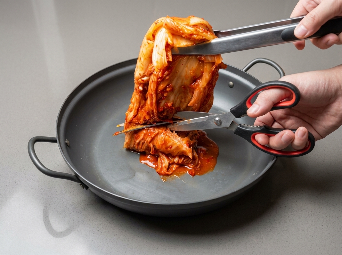 Kitchen scissors cutting whole kimchi into smaller pieces inside a wide cooking pan.