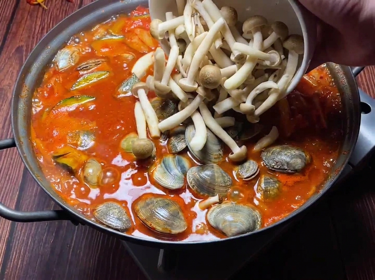 Fresh clams and assorted mushrooms being added to a bubbling red kimchi stew.