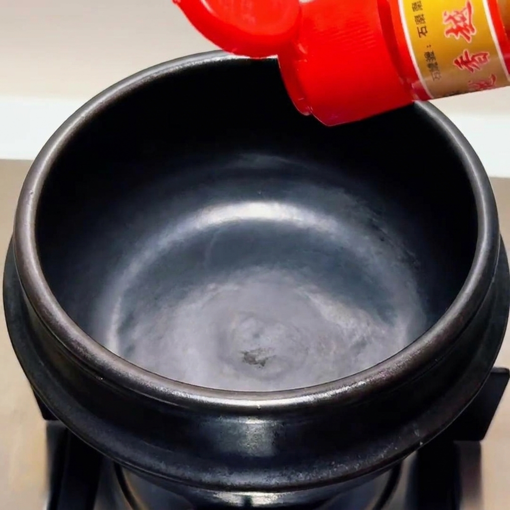Sesame oil being poured from a red-capped bottle into an empty black stone pot.