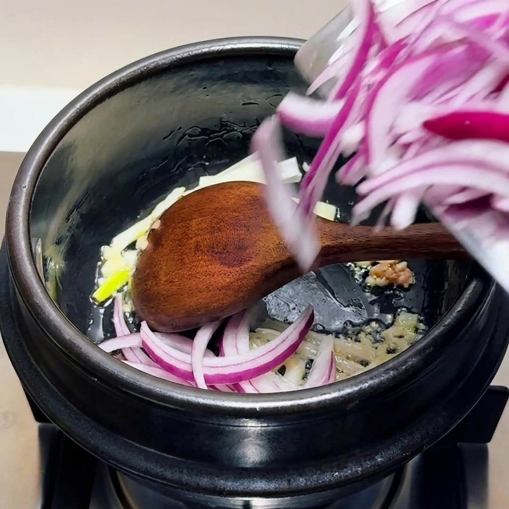 Sliced red onions, minced garlic, and scallion whites being stirred in a black pot with a wooden spoon.