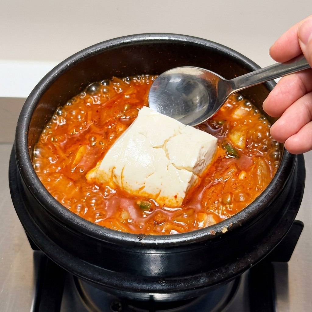 Large chunks of soft silken tofu being carefully scooped into the boiling kimchi stew.