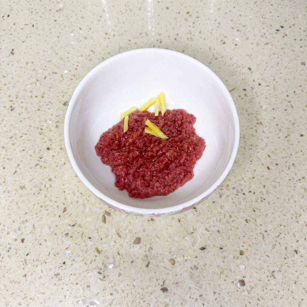 Minced beef in a white bowl mixed with soy sauce, cooking oil, and cornstarch, garnished with ginger slices.