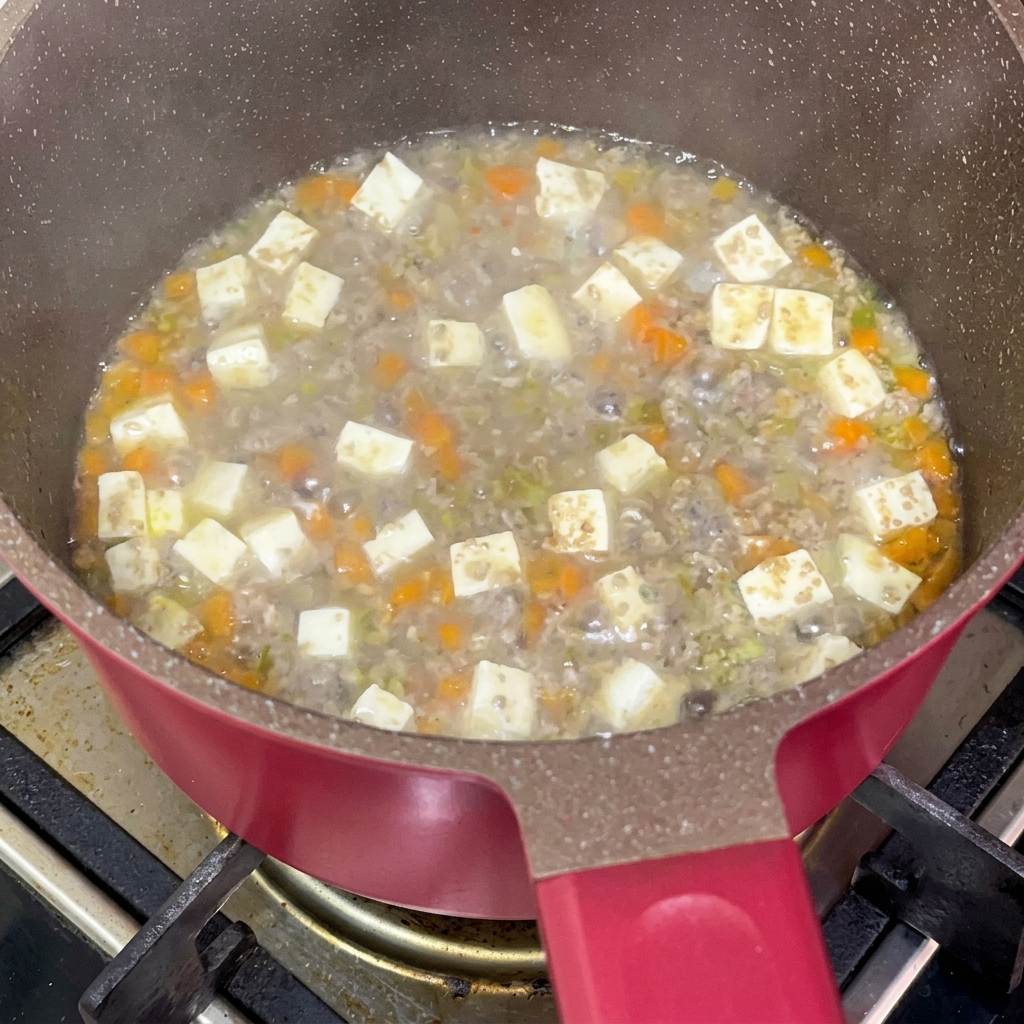 Soy sauce being added to a simmering mixture of minced beef, tofu, and carrots in a pan.