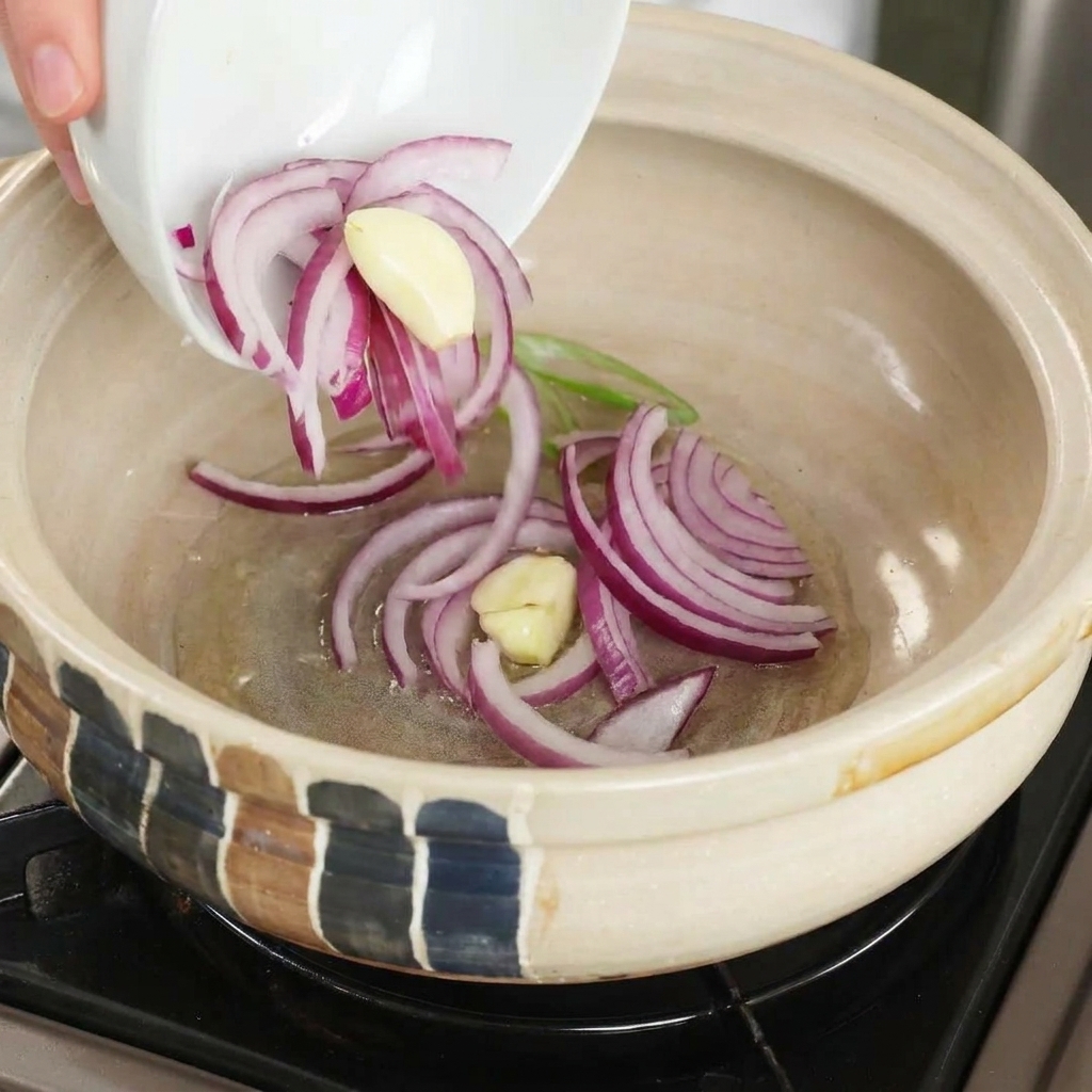 Sliced red onions and garlic cloves being dropped into a heated clay pot with cooking oil.