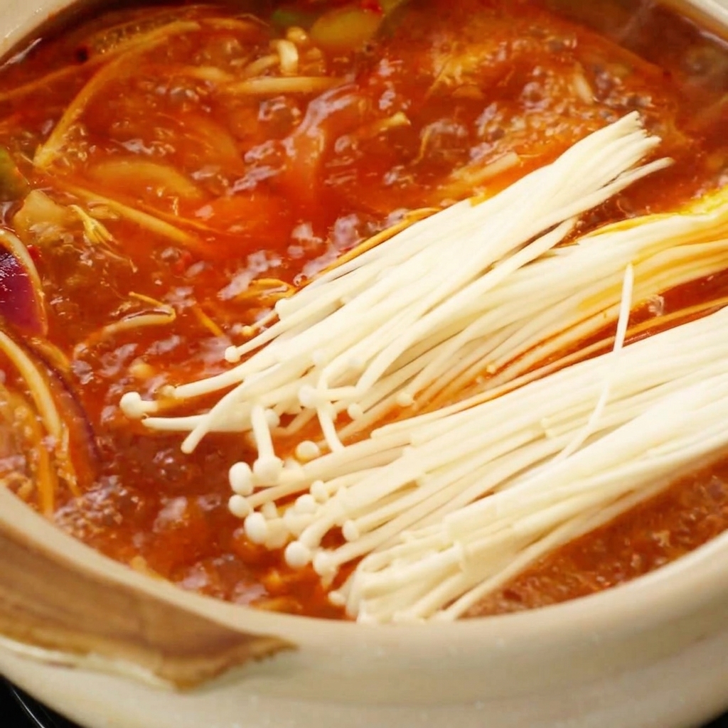 Fresh enoki mushrooms placed into a bubbling red, spicy broth in a clay pot.