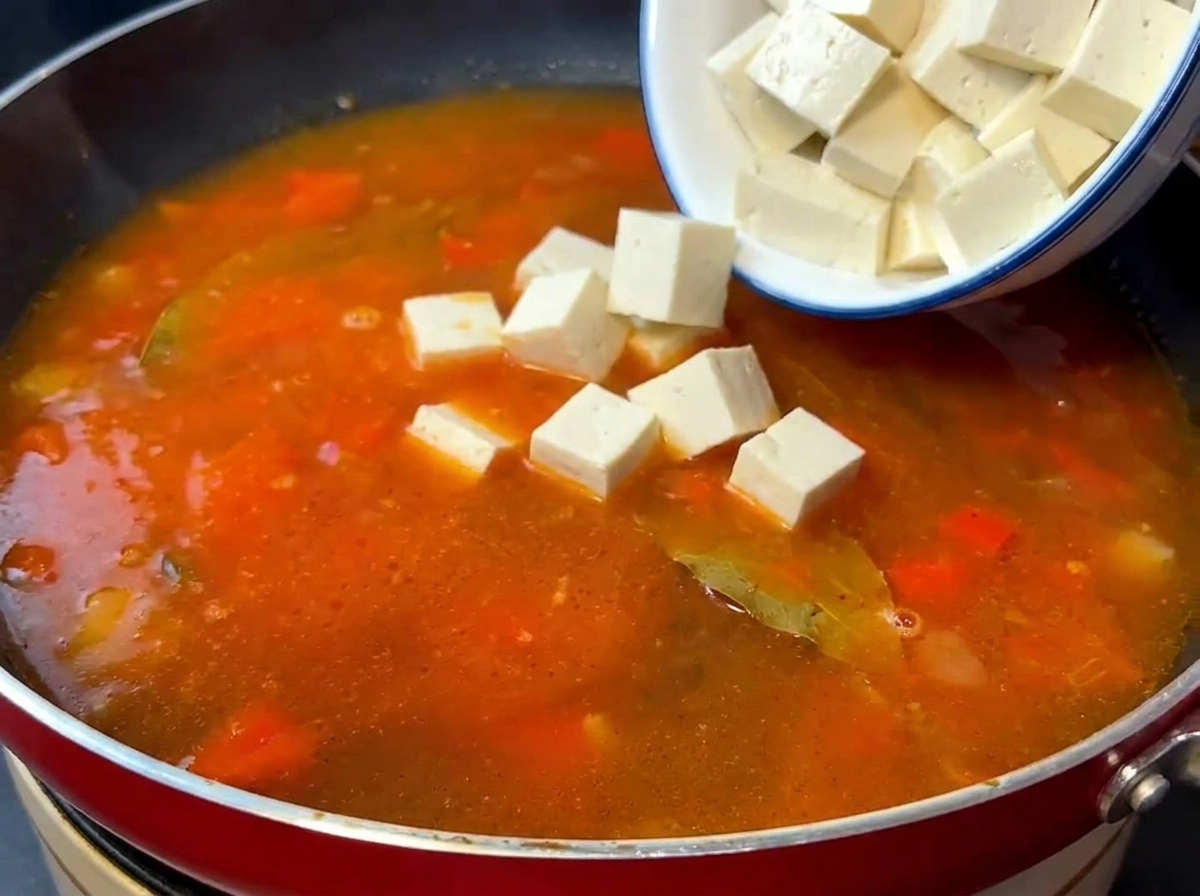 Silken tofu cubes and stir-fried beef being added to a simmering tomato soup base.