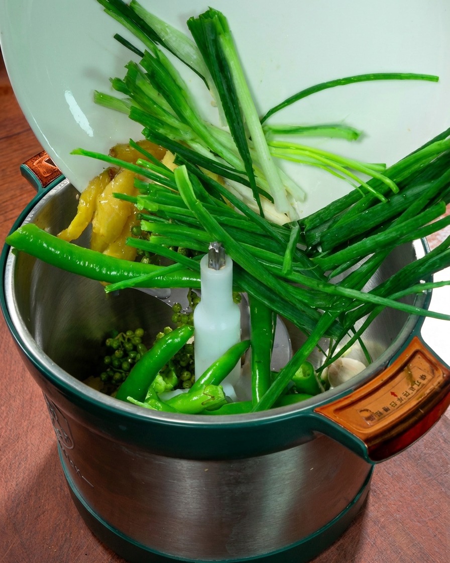 A food processor container filled with green onions, ginger, garlic, fresh green chilies, and Sichuan peppercorns.