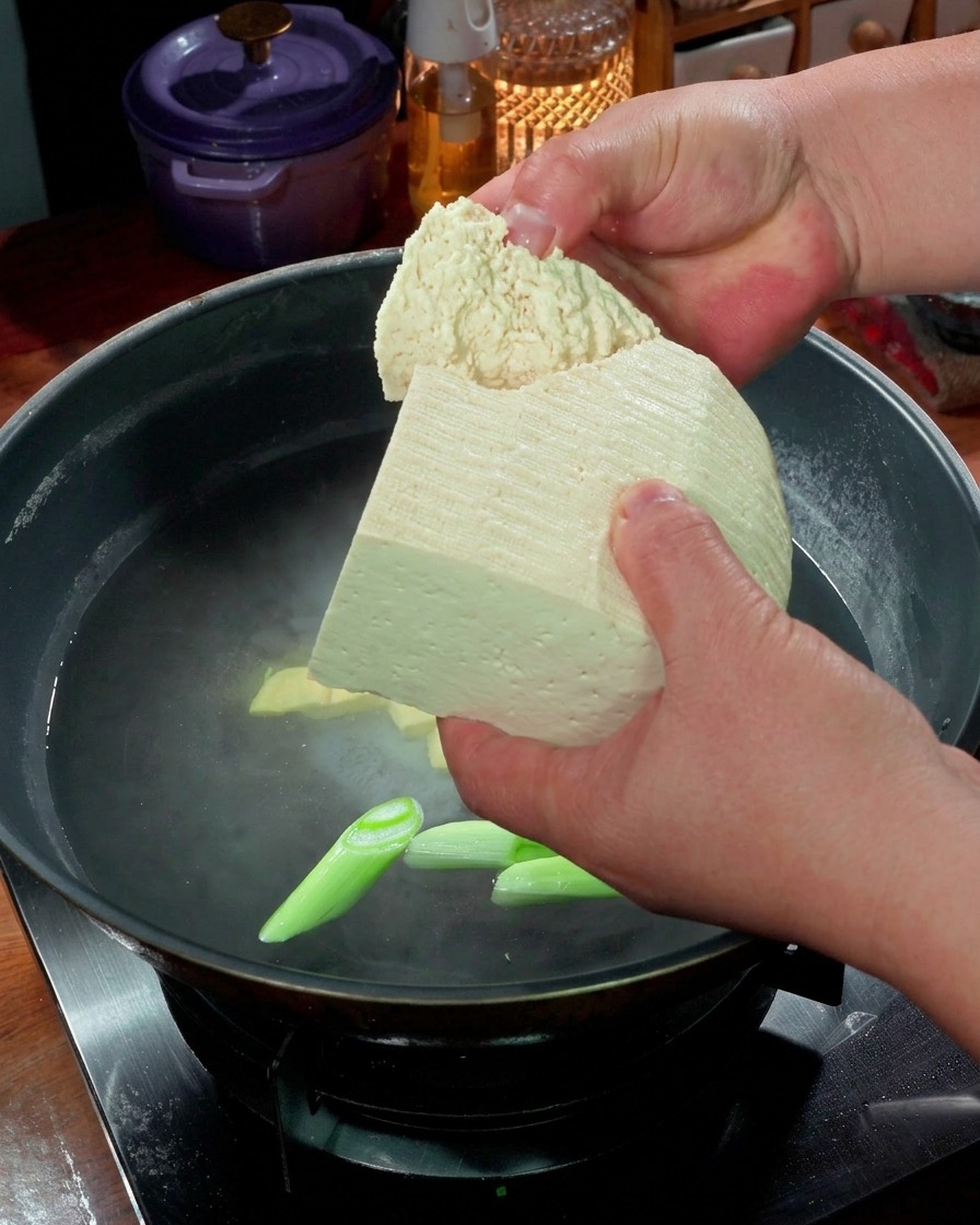 Hands breaking large chunks of firm tofu into a pot of simmering water with green onions.