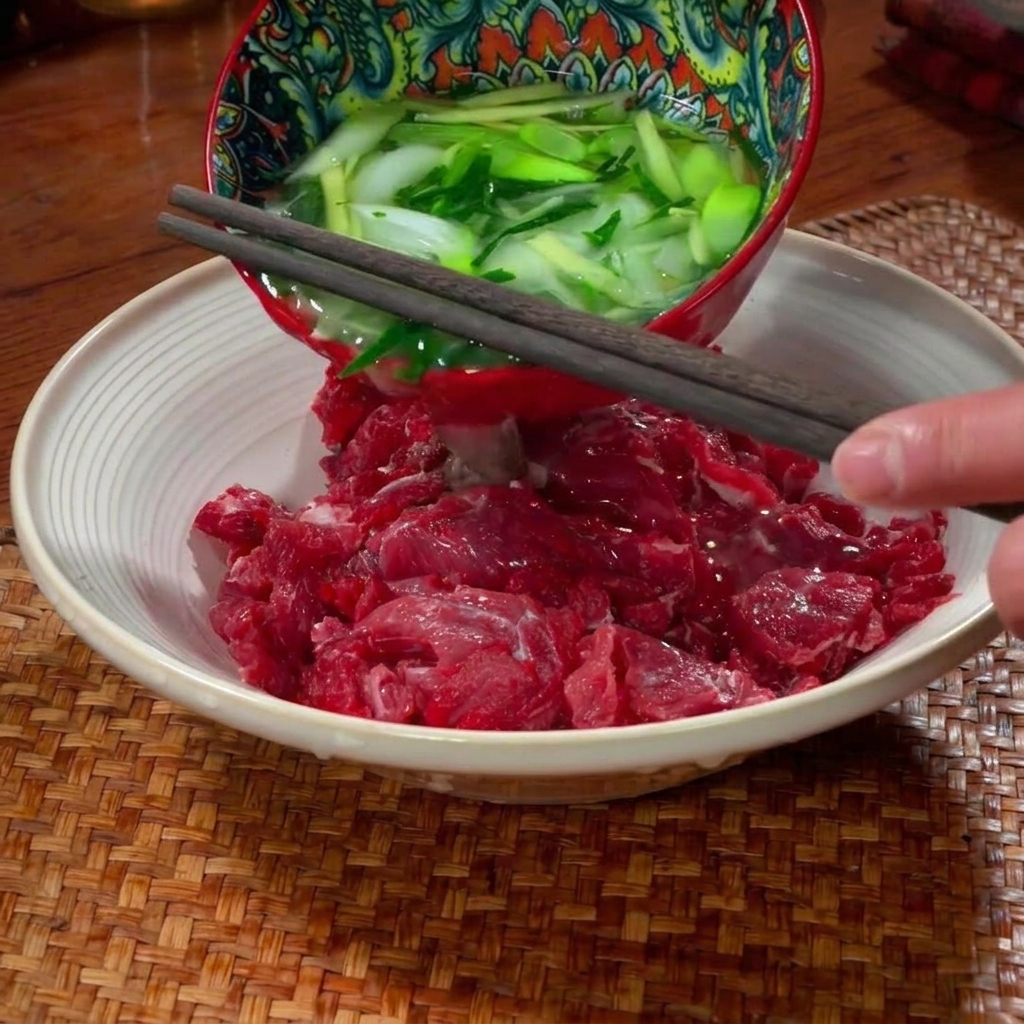 Thinly sliced raw beef being seasoned and massaged by hand in a ceramic bowl.