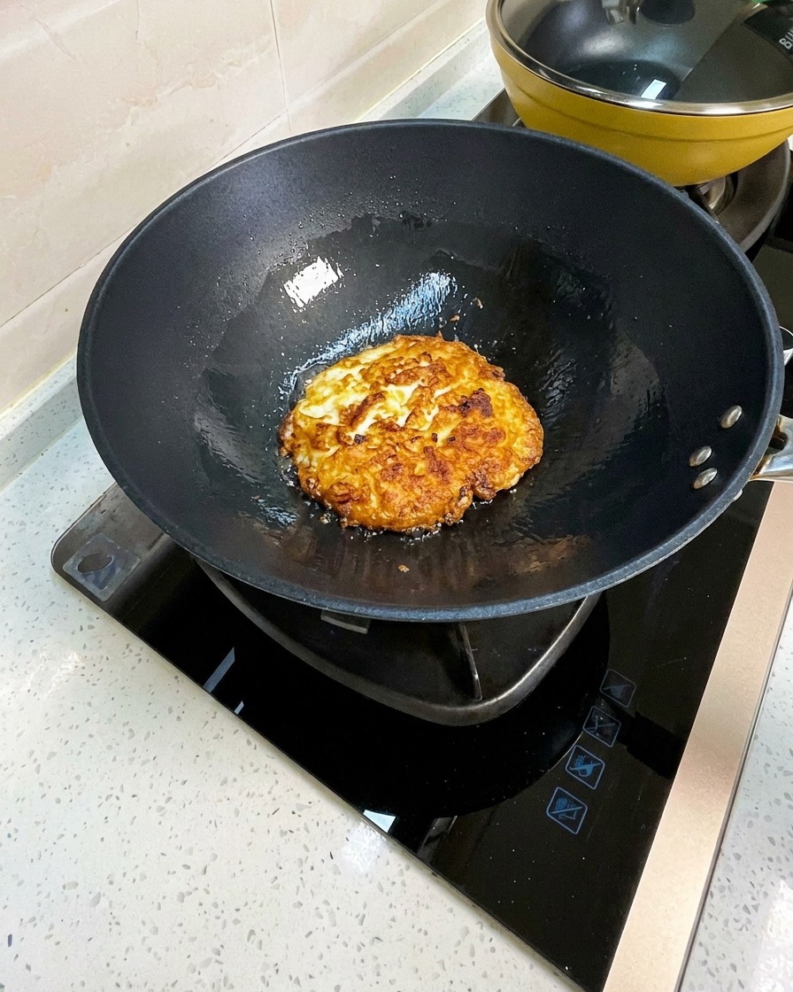 Fried eggs in a wok being broken into large pieces with a metal spatula.