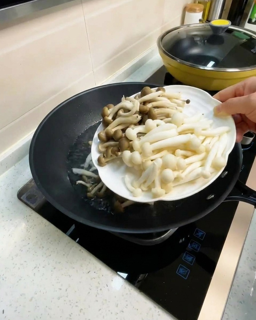 A plate of white beech and crab-flavored mushrooms being added to a wok with boiling broth.