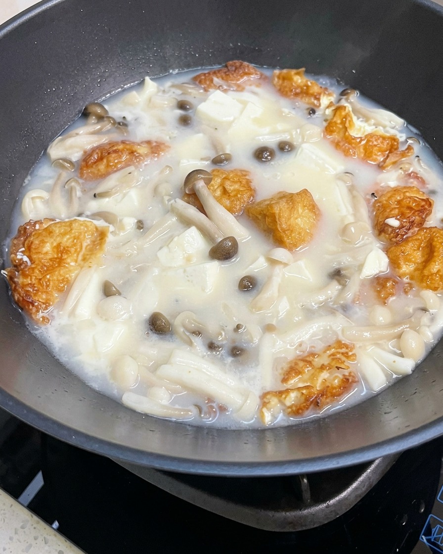 A close-up of the simmering mushroom and tofu soup in a pan being seasoned with salt and pepper.