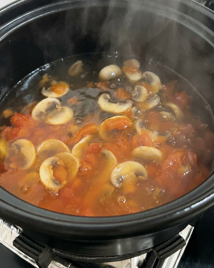 Boiling water being poured into a pot filled with sautéed tomatoes and mushrooms, creating steam.
