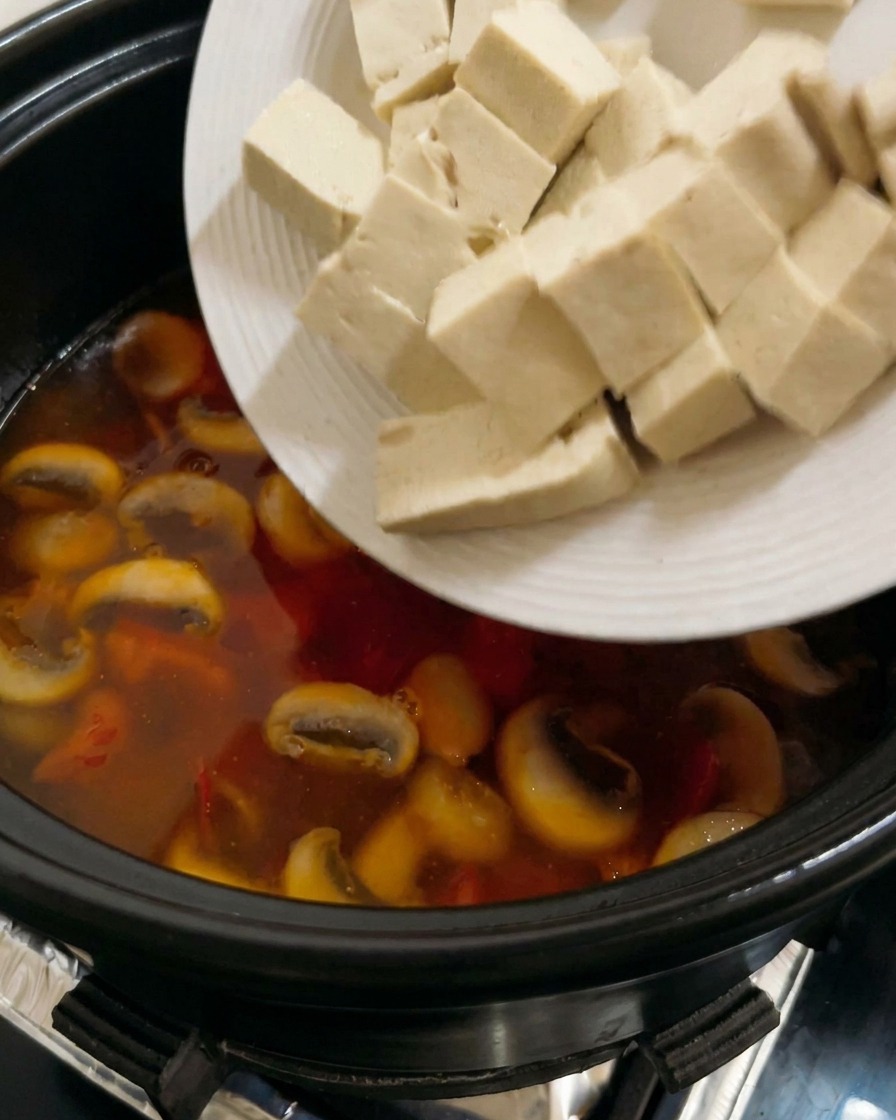 Cubes of firm white tofu being added to a bubbling, tomato-based mushroom soup.