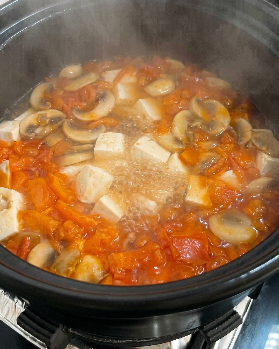 A pot of simmering tomato and mushroom soup with cubed tofu, having beaten egg drizzled into it.