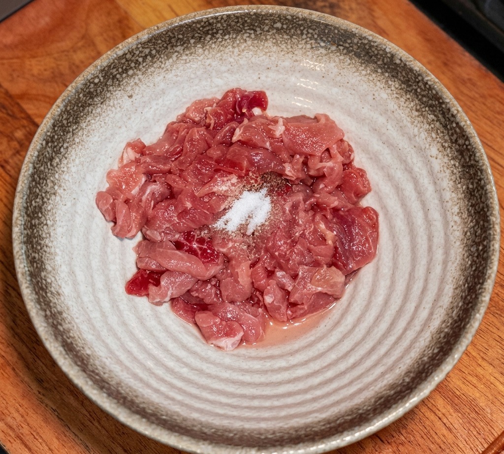 Raw meat strips in a bowl with various seasonings and starch sprinkled on top.