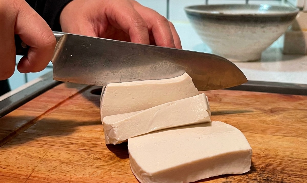 A block of white tofu being sliced into smaller cubes on a wooden board.