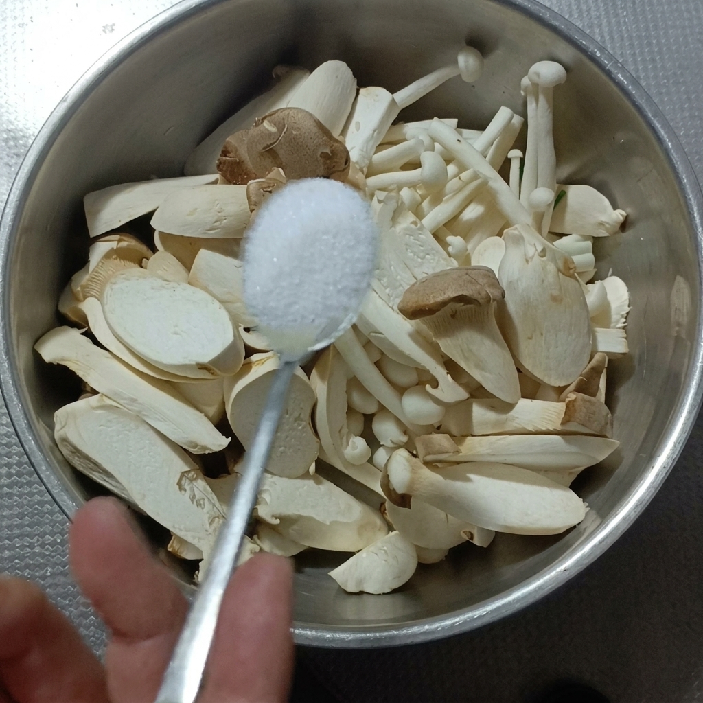 King oyster and white beech mushrooms being salted in a metal bowl.