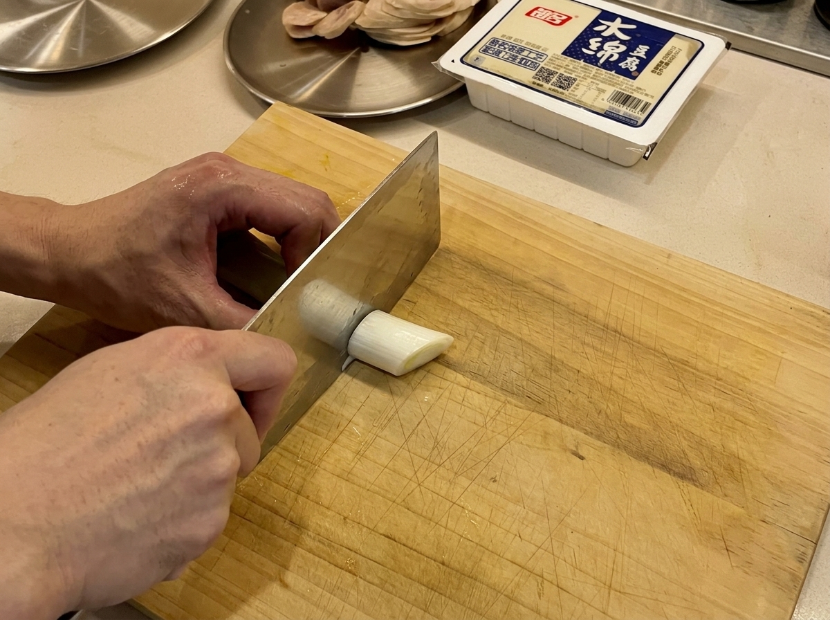 Fresh green scallions being sliced on a wooden board with a cleaver.