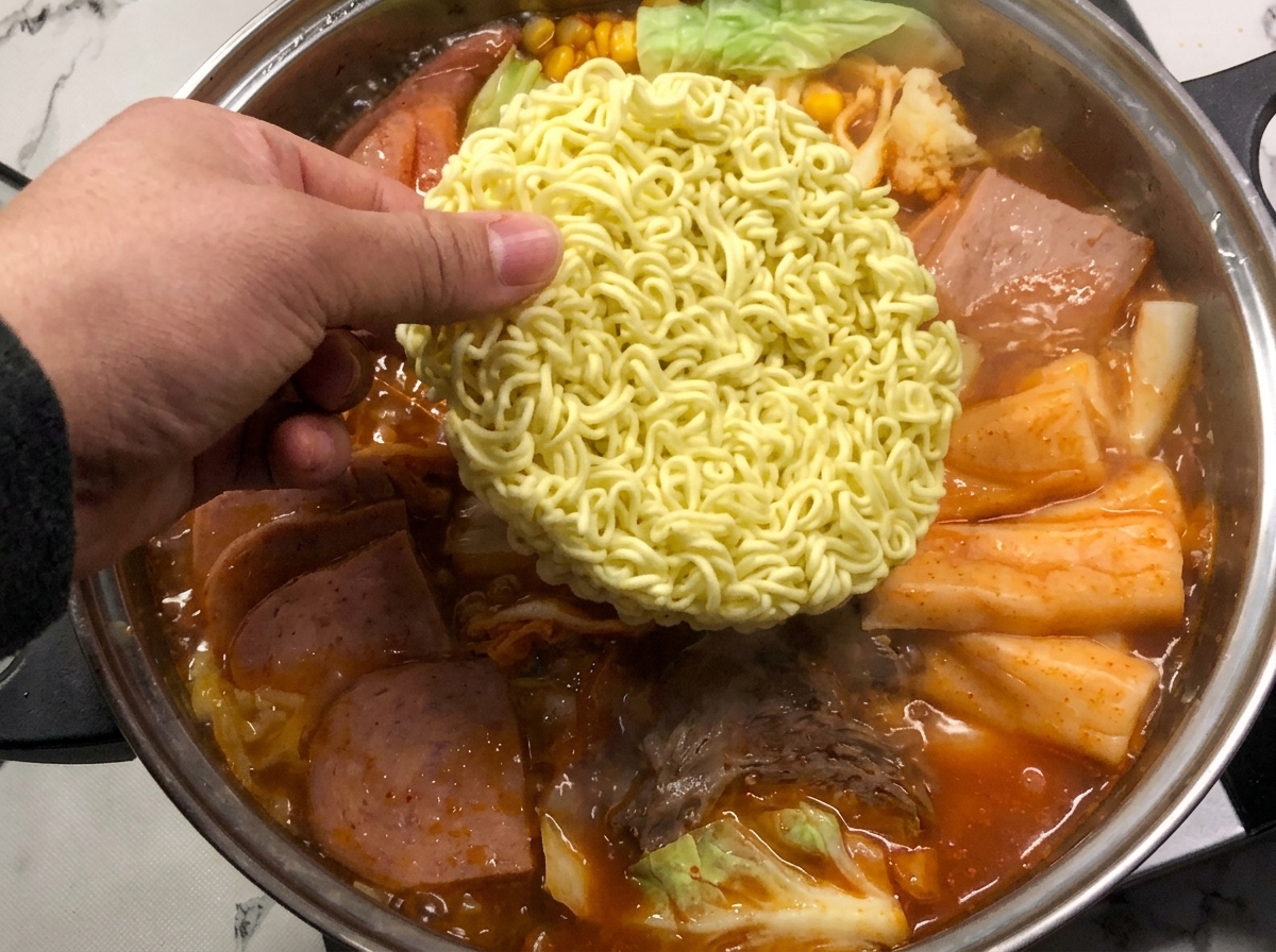 A block of instant noodles being lowered into the vibrant red bubbling broth of the army stew.