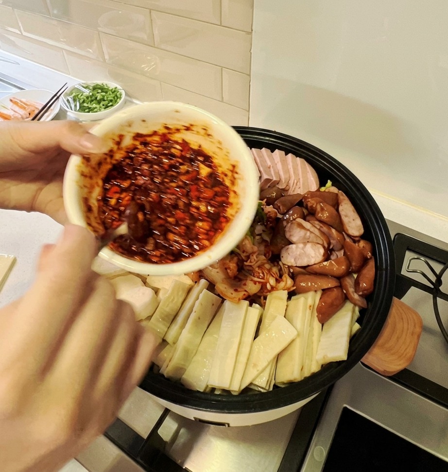 A bowl of vibrant red spicy sauce being poured over the colorful arrangement of ingredients in a large Korean Army Stew pot.