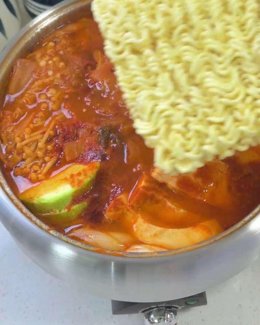 A nearly finished Korean army stew topped with ramen noodles, slices of yellow cheese, and an egg, ready for final cooking.