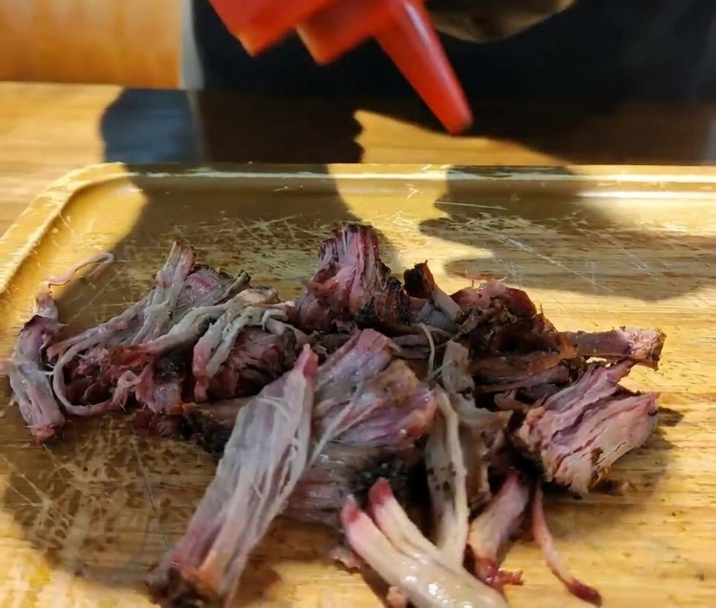A red squeeze bottle pouring thick barbecue sauce onto a pile of shredded smoked pork on a wooden board.