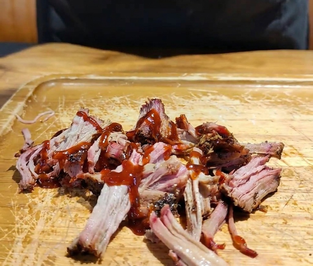 A hand adding a mixture of shredded purple and white cabbage onto a pile of sauced pulled pork on a wooden board.