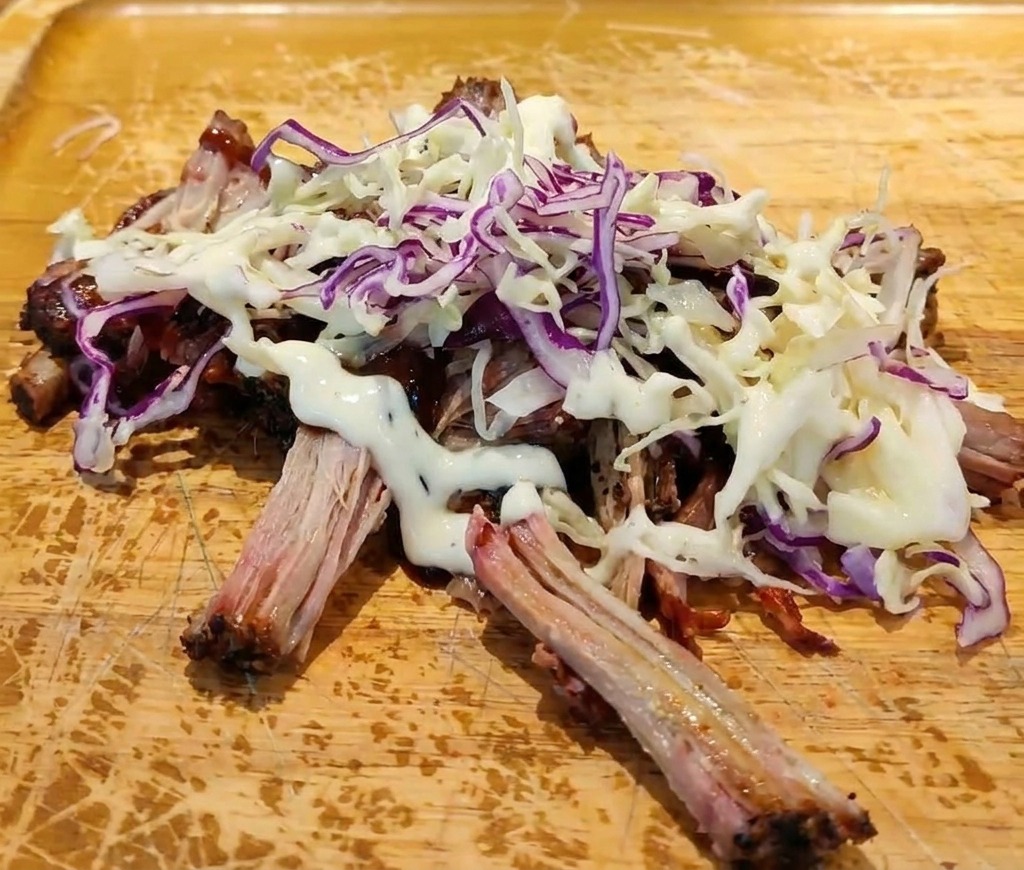 Shredded pork with BBQ sauce topped with a heap of fresh coleslaw and a drizzle of creamy white dressing on a cutting board.