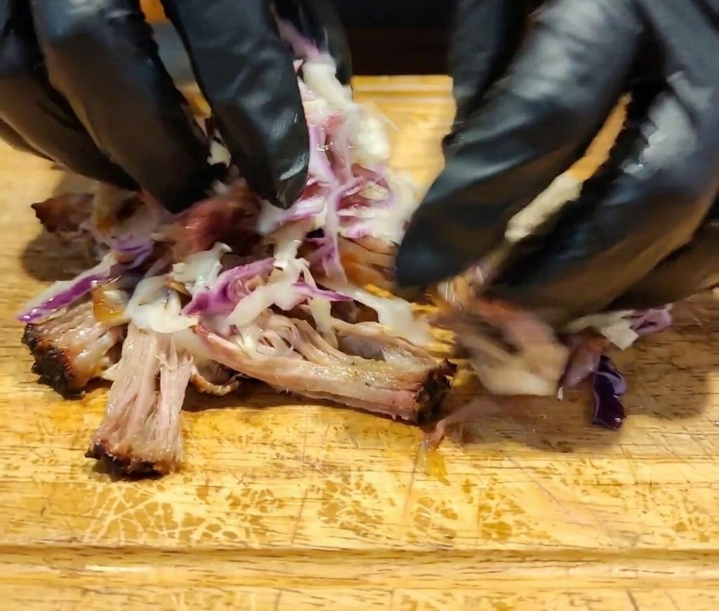 Gloved hands tossing together shredded meat, dark sauce, and colorful coleslaw on a wooden surface.